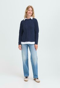 Navyfarbenes Kabelstrickpullover mit Rundhalsausschnitt, kombiniert mit einem weißen Hemd mit Kragen, hellblauen geraden Jeans und beigen Schuhen.