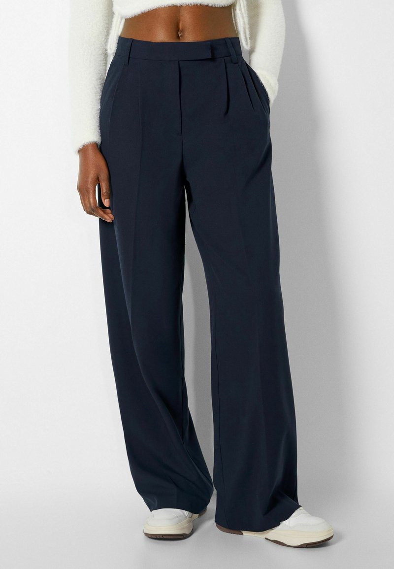 Pantalon large taille haute bleu marine à plis, porté avec des baskets blanches et un pull court blanc pelucheux, mains dans les poches.