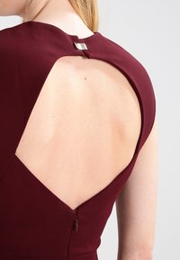 Bordeaux jurk met een uitgesneden achterkant, voorzien van een klein zilveren hardware accent en een verborgen ritssluiting op de onderrug.