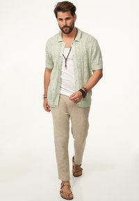 Hellgrünes gestricktes kurzärmliges Shirt mit Knopfleiste, kombiniert mit beigen Leinenhosen und braunen Sandalen mit Riemen. Braune Perlenaccessoires.