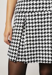 Harmaa ja valkoinen houndstooth-kuvioinen hame, jossa on laskostettu muotoilu, valmistettu sileästä kankaasta, esiteltynä yhdessä käden ja sukkahousujen kanssa.