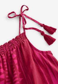Vêtement à motifs rouges avec des bretelles spaghetti dotées de bouts noués et de pompons. Le matériau semble léger avec un corsage smocké.