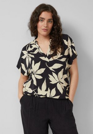 Kobieta o kręconych włosach, ubrana w krótką bluzkę w czarno-biały kwiatowy wzór oraz czarne spodnie, stojąca z rękami w kieszeniach na jednolitym tle.