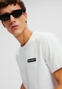 Tricou alb din bumbac cu guler rotund, având o etichetă de marcă neagră pe pieptul stâng. Modelul poartă ochelari de soare negri, cu un design unghiular.