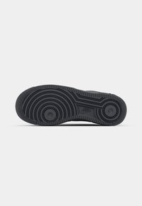 Semelle de chaussure de sport en caoutchouc noir avec des motifs circulaires et un petit logo Nike près de la voûte plantaire, présentée sur un fond blanc uni.