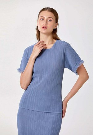 Femme portant un haut bleu côtelé à manches courtes et une jupe assortie, posant avec une main touchant son cou et l’autre sur sa hanche.