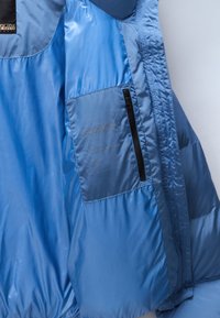 Interno di un giubbotto puffer azzurro chiaro, caratterizzato da un tessuto in nylon liscio, una tasca con zip e un dettaglio del logo in rilievo nell'area della tasca.