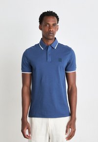 Blå polo skjorte med hvid trim, der har en to-knap lukning, korte ærmer og et diskret mærke logo på brystet. Glat bomuldsstruktur.
