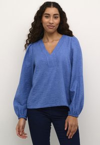 Blouse bleu, texturée, à manches longues avec un décolleté en V et des poignets froncés. Assortie à un jean foncé, mettant en avant une coupe décontractée et un style casual.