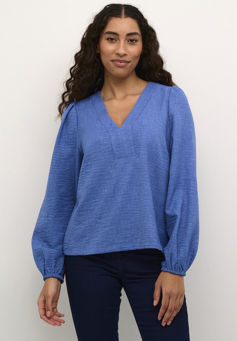 Blouse bleu, texturée, à manches longues avec un décolleté en V et des poignets froncés. Assortie à un jean foncé, mettant en avant une coupe décontractée et un style casual.