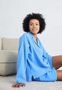 Set de pijama din bumbac de culoare bleu deschis, cu cămașă cu nasturi și pantaloni scurți, având o croială lejeră și o țesătură moale, texturată. Design simplu, fără modele.