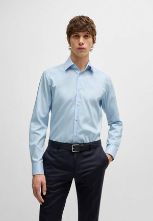 Chemise boutonnée à manches longues bleu clair avec un col évasé, tissu lisse et éclat subtil, associée à un pantalon sombre et une ceinture noire.