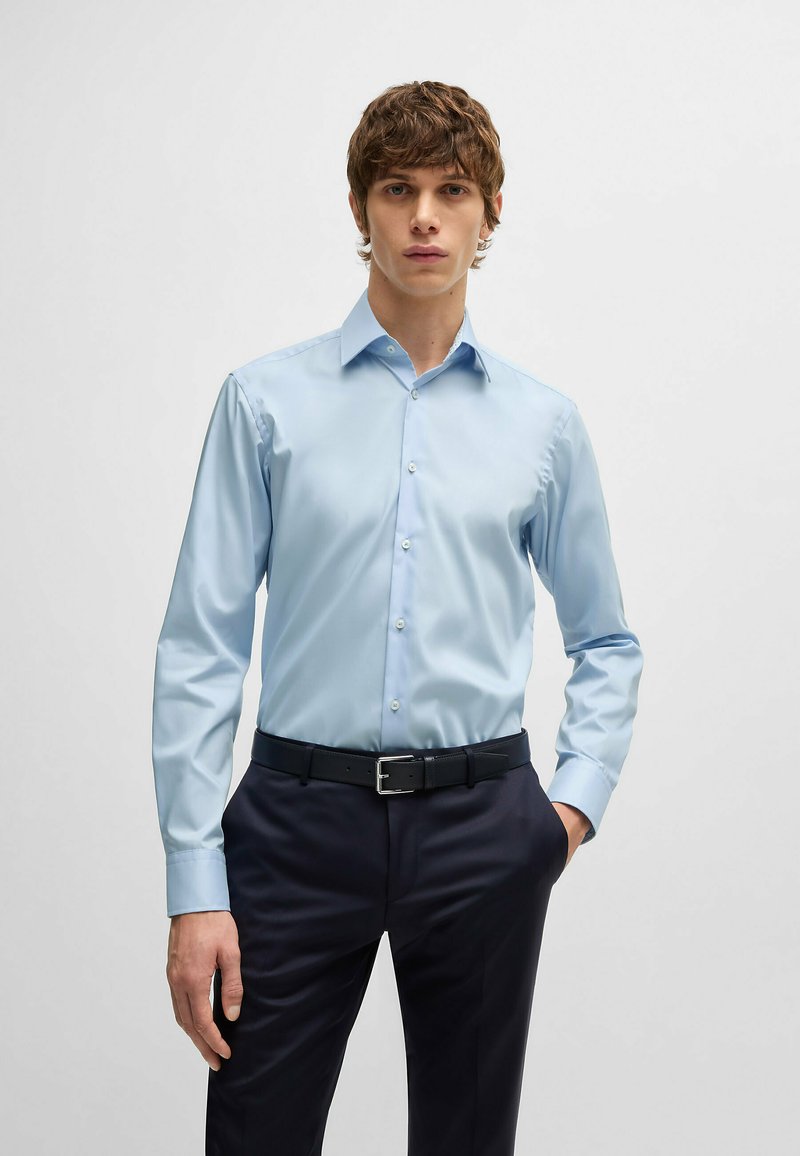 Chemise boutonnée à manches longues bleu clair avec un col évasé, tissu lisse et éclat subtil, associée à un pantalon sombre et une ceinture noire.