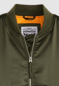 Olive green bomber jacket with a silver zipper, ribbed collar, and orange lining. Features a branded tag inside the collar.