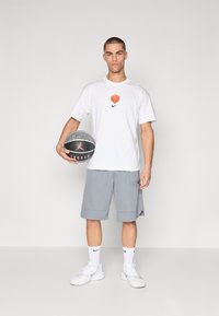 Biela Nike tričko s oranžovým logom, šedé športové šortky, biele tenisky a čierny basketbal s červeným logom, umiestnené na jednoduchom pozadí.