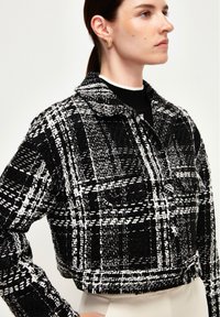 Chaqueta de cuadros en blanco y negro, recortada, con un tejido texturizado, cuello de camisa y bolsillos frontales, combinada con una blusa negra debajo.