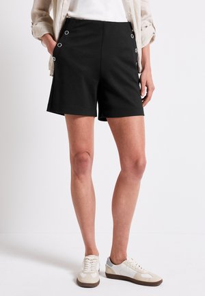 Shorts noirs avec poches latérales et accroches en œillets argentés. Tissu doux, coupe droite et longueur mi-cuisse. Associés à des baskets blanches.