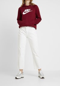 Vinröd hoodie med vitt Nike-logotyp, kombinerad med vita högmidjade jeans och grå sneakers. Har en framficka och huva med dragsko.