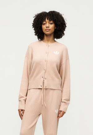 Vrouw met krullend haar die een beige knoopvest draagt en bijpassende broek met trekkoorden met witte Adidas-logo's op borst en dij.