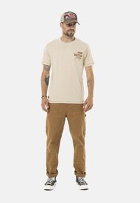 Camiseta de algodón beige con estampado rojo en el pecho izquierdo, combinada con pantalones cargo marrones y zapatillas de deporte en blanco y negro. La gorra es gris y está detallada.