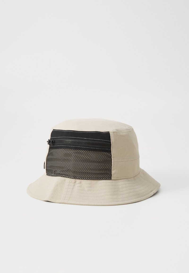 Bucket hat in beige cotton with a black mesh side panel; features a zippered pocket across the top section. Soft, structured shape.