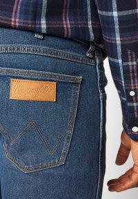 Mėlyni džinsai su trikampiu siuvinėjimo raštu ant galinio kišenės; turi odinį Wrangler logotipą ir tiesų siluetą.