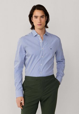 Joven con cabello oscuro de longitud media, vistiendo una camisa de botones a rayas azules y blancas y pantalones verde oscuro, de pie contra un fondo neutro.