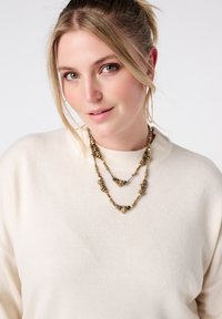 Gouden gelaagde halsketting met gestructureerde kralen en natuurlijke accenten, gedragen over een lichtbeige trui met een ronde hals.