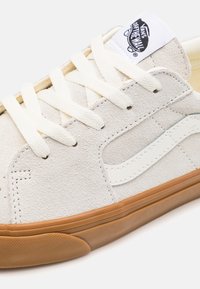 Weißer Sneaker aus Wildleder mit cremefarbenen Schnürsenkeln, Gummisohle, blauen Akzenten und einem Vans-Logo-Label auf der Zunge. Verfügt über ein klassisches Low-Top-Design.