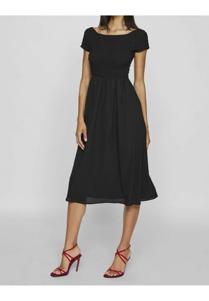 Femme portant une robe midi noire à épaules dénudées et des sandales rouges à talons hauts avec des brides, debout devant un fond gris uni.