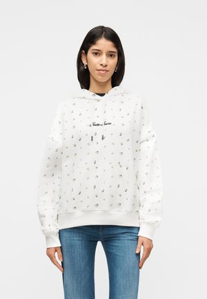 Γυναίκα που φοράει λευκή φούτερ με κουκούλα με μικρά λουλουδάτα σχέδια και το κείμενο "Guess Jeans", συνδυασμένη με μπλε τζιν, στέκεται μπροστά σε απλό φόντο.