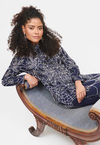 Námořnický modrý šaty s propracovaným stříbrným korálkovým zdobením. Dlouhé rukávy a vysoký límec. Model sedí na zdobené dřevěné pohovce.