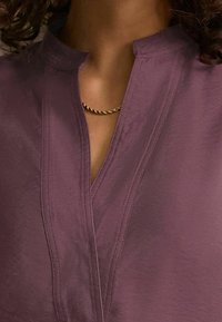 Blusa viola con collare alla mandarina e apertura a chiave, caratterizzata da dettagli di cucitura sottili e una texture liscia. Accento con collana in catena dorata.