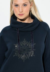 Sudadera azul marino con cuello de cisne, que presenta un patrón metálico de copos de nieve en la parte delantera y cordones de tela con puntas metálicas.