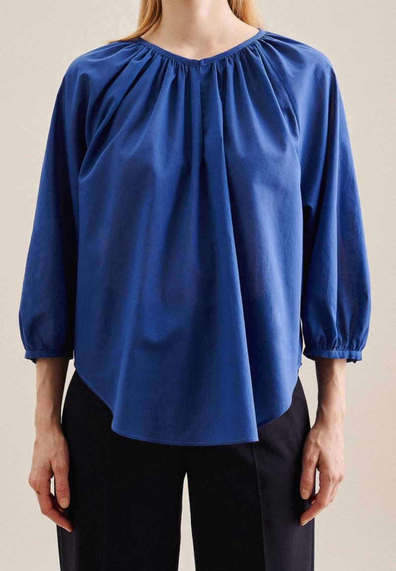 Blusa di cotone blu con scollatura arricciata, maniche a tre quarti e vestibilità morbida, caratterizzata da un orlo arrotondato e una superficie liscia.