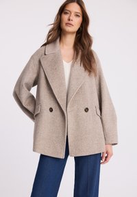 Beżowy, dwurzędowy płaszcz z wełny z wyciętym kołnierzem, w oversize'owym fasonie, z dwiema przednimi kieszeniami, zestawiony z ciemnoniebieskimi spodniami o szerokich nogawkach.