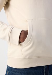 Felpa beige con tasca a marsupio, polsini e orlo a coste. Tessuto liscio, forma aderente, abbinata a jeans blu denim.