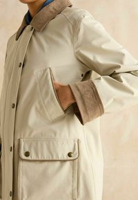 Jachetă impermeabilă bej deschis, cu un guler din catifea brună, nasturi tip clapetă și o buzunar frontal, având o textură netedă și o croială lejeră.