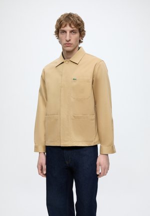 Man with light brown curly hair wearing a beige jacket with front pockets and dark blue jeans against a plain white background.