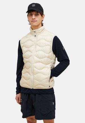Beige puffer vest met quiltpatroon, ritssluiting aan de voorkant, staande kraag en zijzakken. Gecombineerd met een marineblauw langemouwen shirt en zwarte shorts.