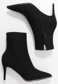 Paire de bottines en suède noir à bout pointu avec talons hauts fins et fermeture éclair latérale sur fond blanc.