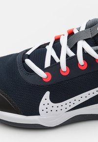Jūras spēku tīkla sporta apavi ar baltām šņorēm, spilgti sarkaniem acs caurumiem un baltu Nike logotipu. Ievērojams melns pirkstu vāciņš un strukturēts materiāls.
