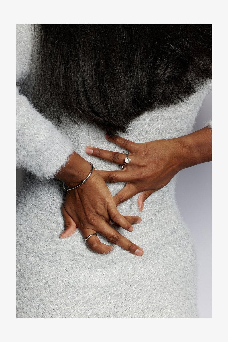 Maglione bianco, morbido e texturizzato, aderente al corpo, con le mani posizionate dietro. Anelli e bracciali adornano i polsi.