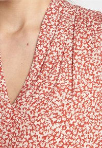 Tessuto rosso con un motivo floreale bianco, caratterizzato da un collo a V e dettagli arricciati, con una texture liscia e leggera.