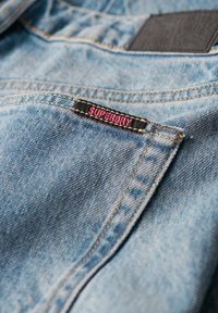 Jean en denim bleu clair avec un tissu texturé, présentant une étiquette en cuir noir et une étiquette rose 'SUPERDRY' sur la poche avant.