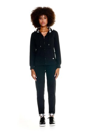 Une femme porte un sweat à capuche noir zippé et un pantalon de jogging assorti avec des bordures à carreaux, devant un fond blanc.