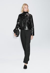 Zwarte leren cropped jack met een glanzende afwerking, gecombineerd met zwarte broek en sandalen. Houdt een zwarte gevlochten clutch vast.