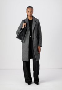 Cappotto grigio in lana con colletto a revers, chiusura con bottoni e taglio dritto, abbinato a pantaloni neri a gamba larga con texture e una borsa nera.