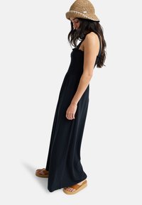 Γυναίκα που φοράει μακρύ μαύρο αμάνικο φόρεμα, ταμπά πλατφόρμες σανδάλια και πλεκτό ταμπά καπέλο τύπου κουβά, στέκεται πλάγια μπροστά σε ένα απλό φόντο.