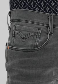 Šedé džínové kalhoty s zadní kapsou ve tvaru písmene V a měděným nýtem. Textura je hladká s jemnými šicím detaily.